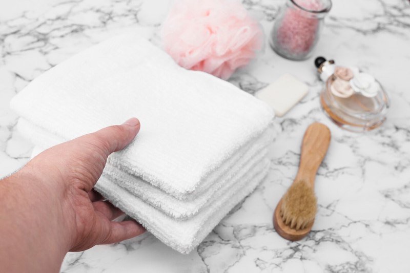 Hand holding folded white towels on a marble surface with bath accessories