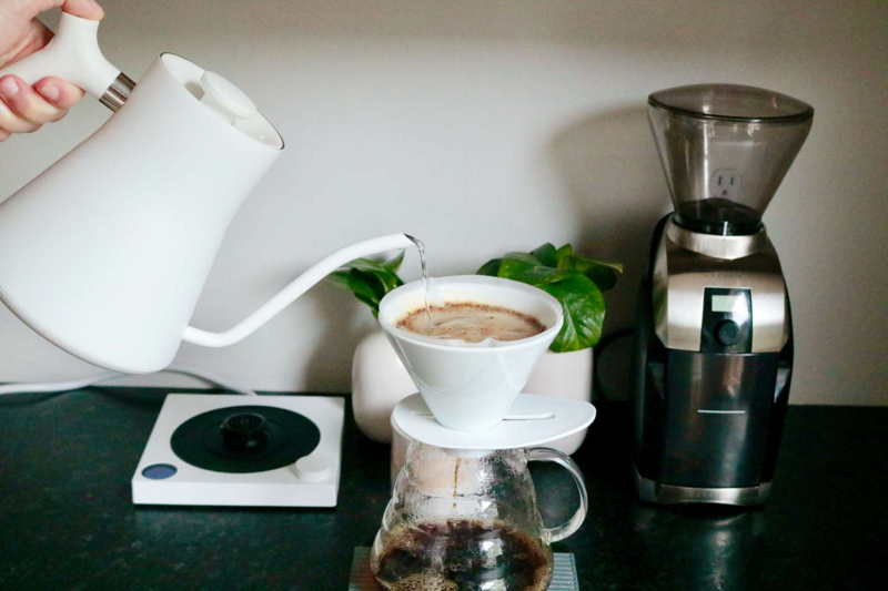 Pour-over coffee setup with white kettle, grinder, and glass carafe on a countertop