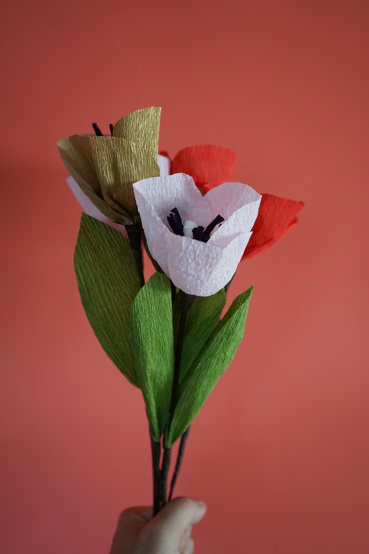 Hand holding colorful paper flowers, red, white, and gold on green stems